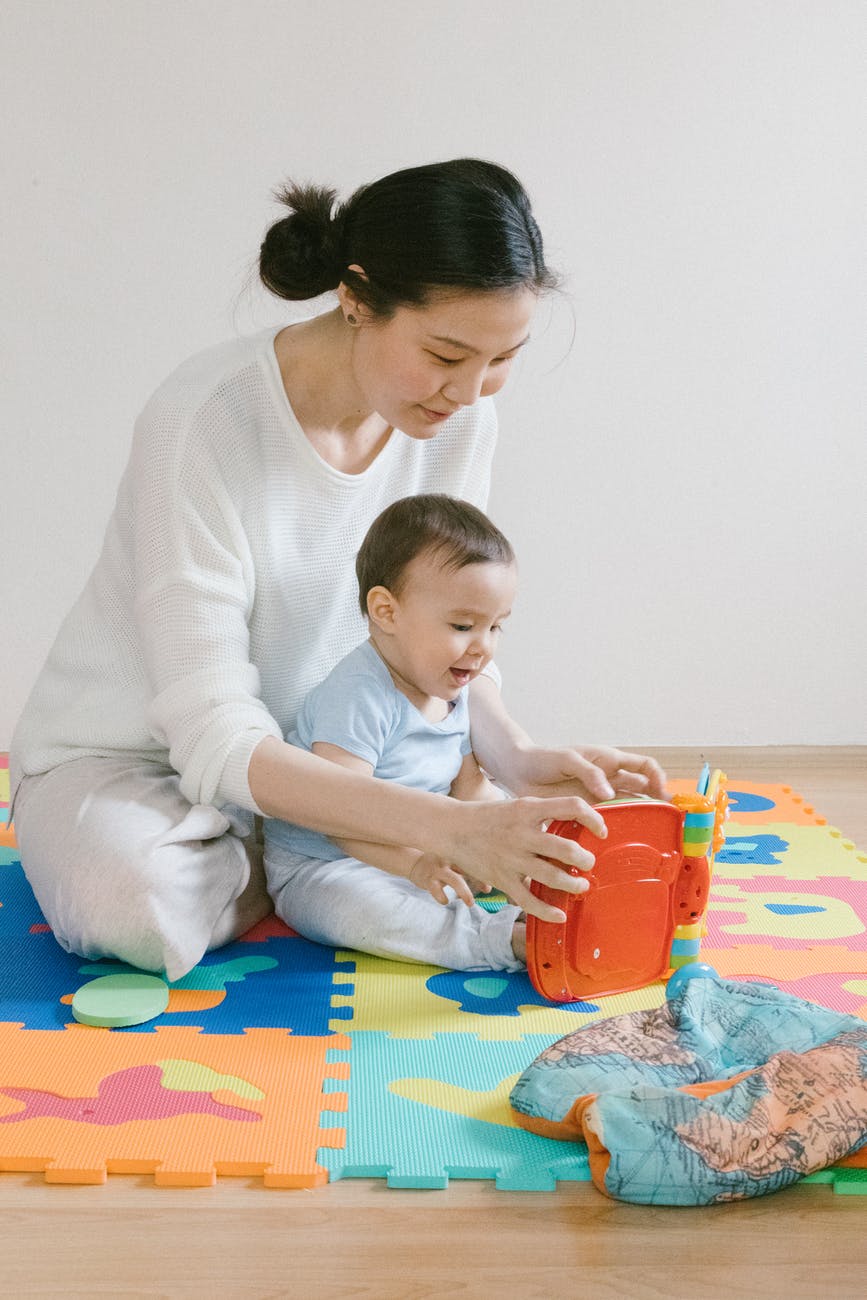 woman in white long sleeve shirt sitting beside a baby playing with the toy
